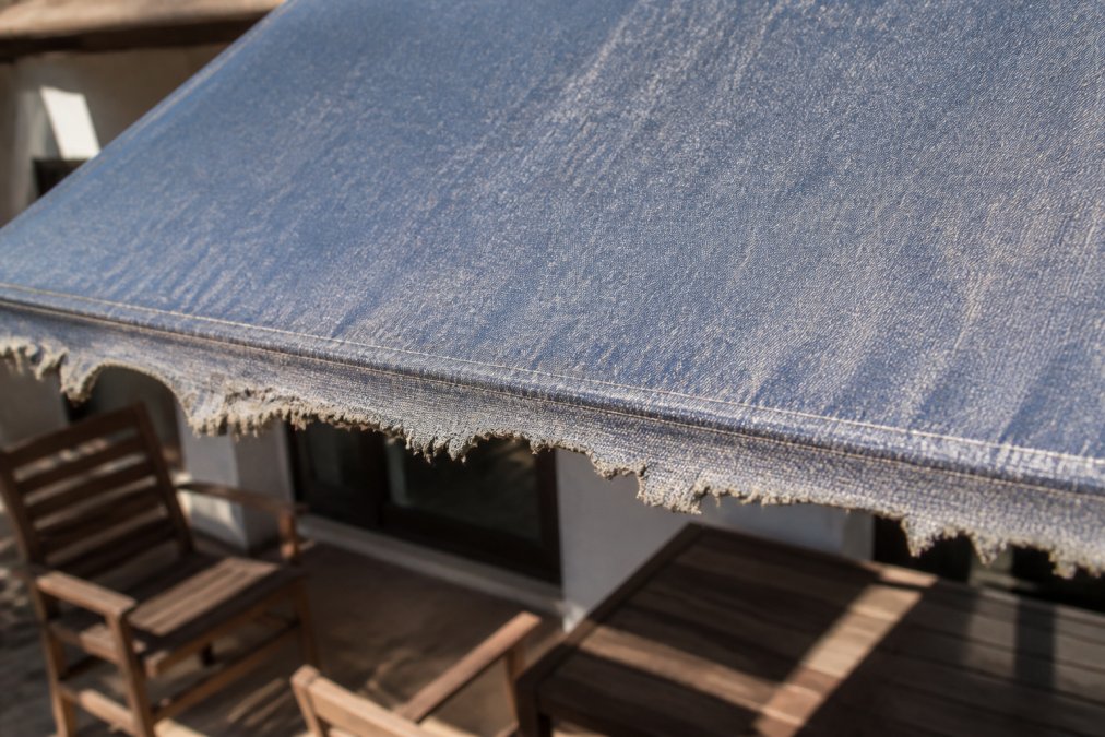Close-up of a sun-damaged awning fabric showing faded color and frayed edges, with bright sunlight casting harsh shadows on a patio