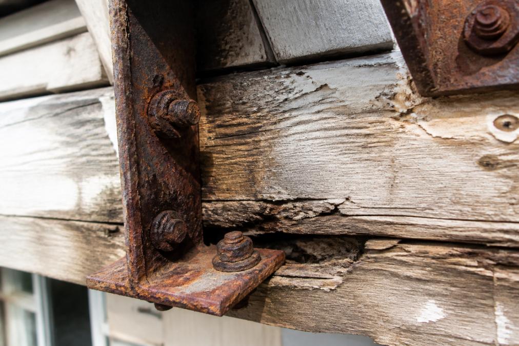 Close-up of heavily corroded metal bolts and brackets on a coastal balcony structure showing rust discoloration and material loss, with weathered wood framing visible in background