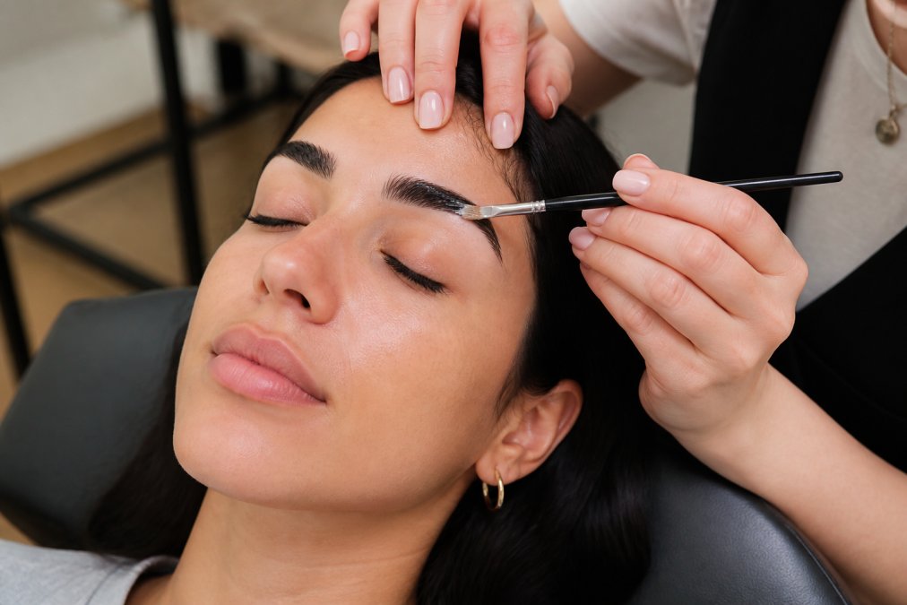 Close-up of a brow artist applying lamination solution to a client's thick, dark eyebrows with a small brush in a bright, modern salon