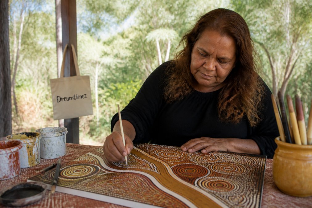 Indigenous artist working on traditional artwork in their studio or outdoor setting, hands visible creating intricate patterns with natural materials, natural daylight illuminating the creative process