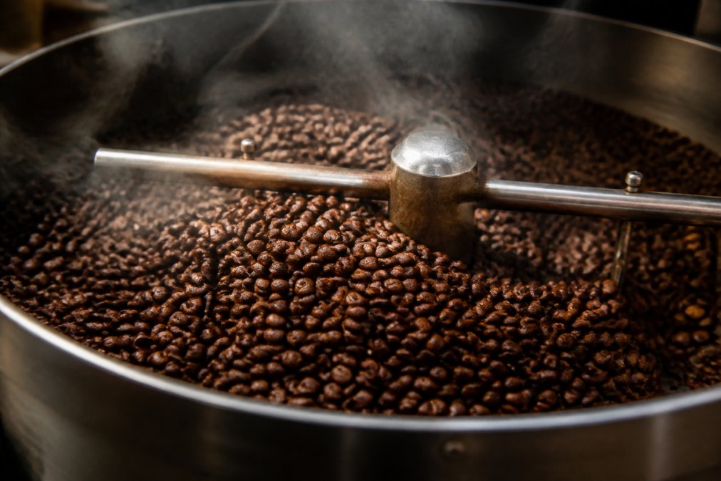 Close-up of freshly roasted coffee beans cooling in a metal roasting drum, wisps of steam rising, showing deep brown color variation and glossy surface in warm industrial lighting
