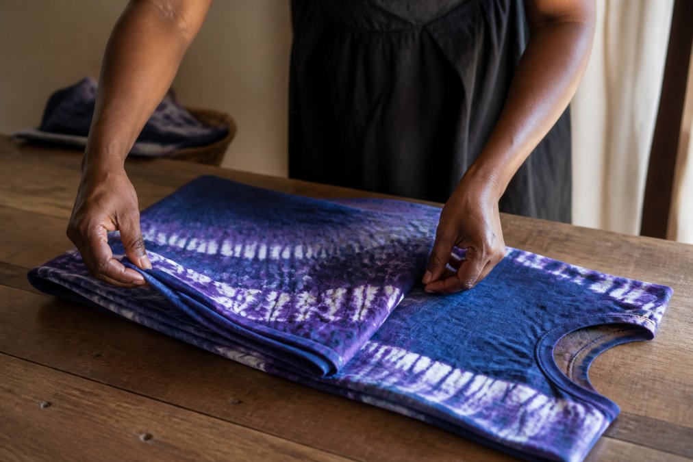 A woman's hands carefully folding a vibrant hand-dyed Adire cotton dress on a wooden table, natural light highlighting the unique gradient patterns and artisanal texture