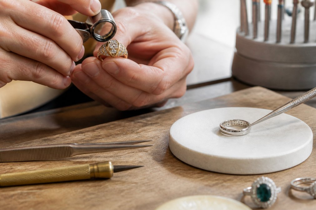 Jeweler's hands examining an antique diamond under magnification, inspecting hallmarks on a platinum band with precision tools under bright studio lighting