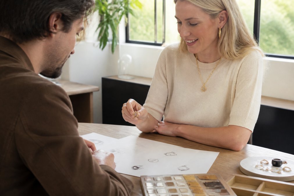 Designer and client reviewing jewelry sketches and metal samples together at a workspace, discussing custom design concepts with natural light from large windows