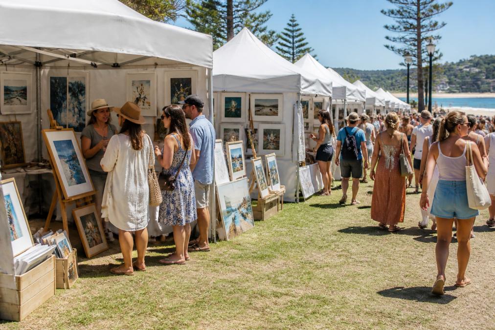 An outdoor art fair in a coastal Australian setting with white stalls displaying original paintings and prints, visitors in casual summer clothing browsing artworks and speaking with artists under a bright midday sun