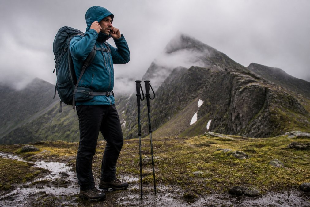 A hiker in a remote mountain landscape adjusting a technical hooded jacket, backpack and trekking poles planted in wet ground, overcast skies and low clouds behind the ridge