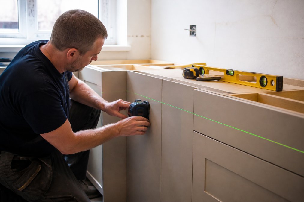 An installer in work clothes positioning a laser level marker on the end panel of a kitchen cabinet run, with a spirit level and tape measure resting on the worktop nearby, daylight coming through a window