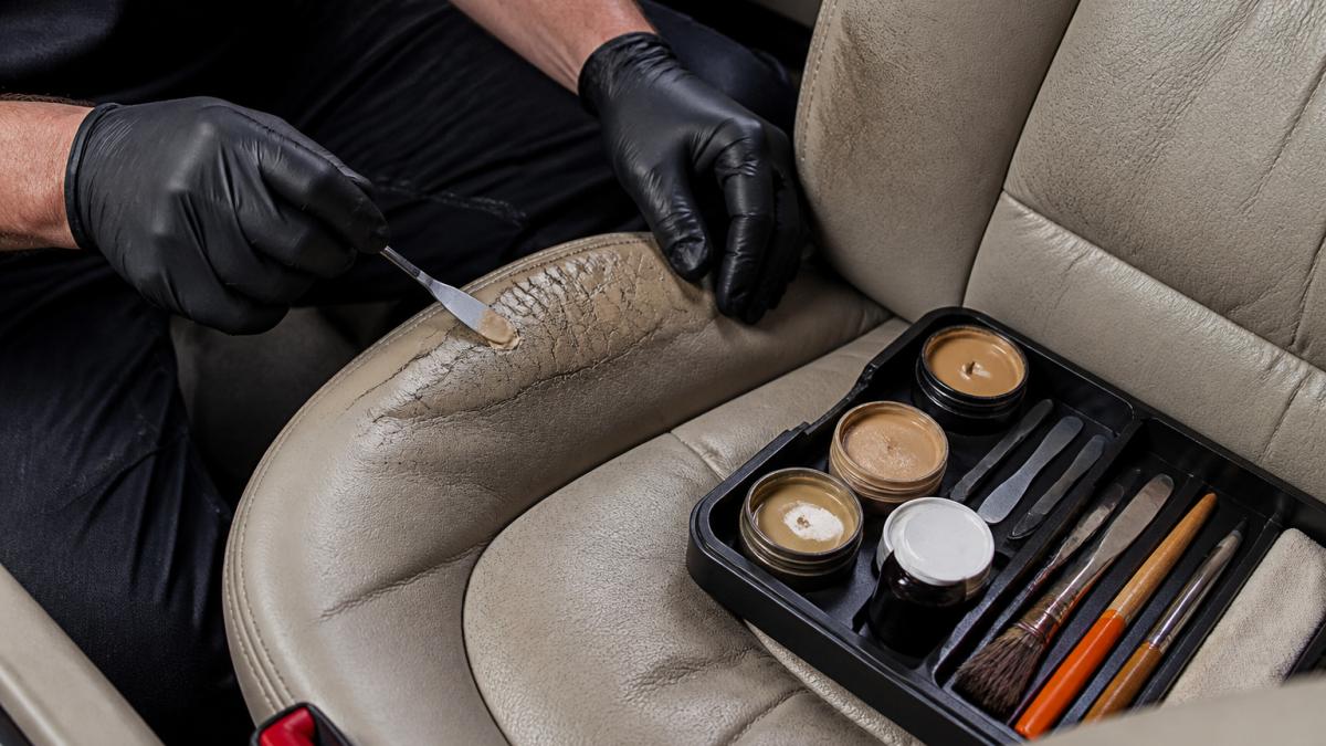 Close-up of a technician's gloved hands applying colour-matched filler to a cracked leather car seat bolster, with a small tool kit resting on the seat beside them