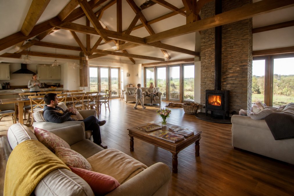 Spacious interior of a converted barn showing exposed wooden beams, high ceilings, comfortable seating arranged around a log burner, large windows with countryside views, and multiple family members relaxing in different areas of the open-plan living space with natural afternoon light streaming in