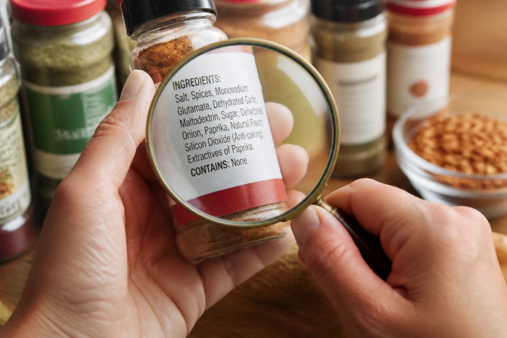 Close-up of hands examining spice jar labels with ingredient lists visible, magnifying glass showing fine print and additives highlighted on a commercial spice container in natural kitchen lighting