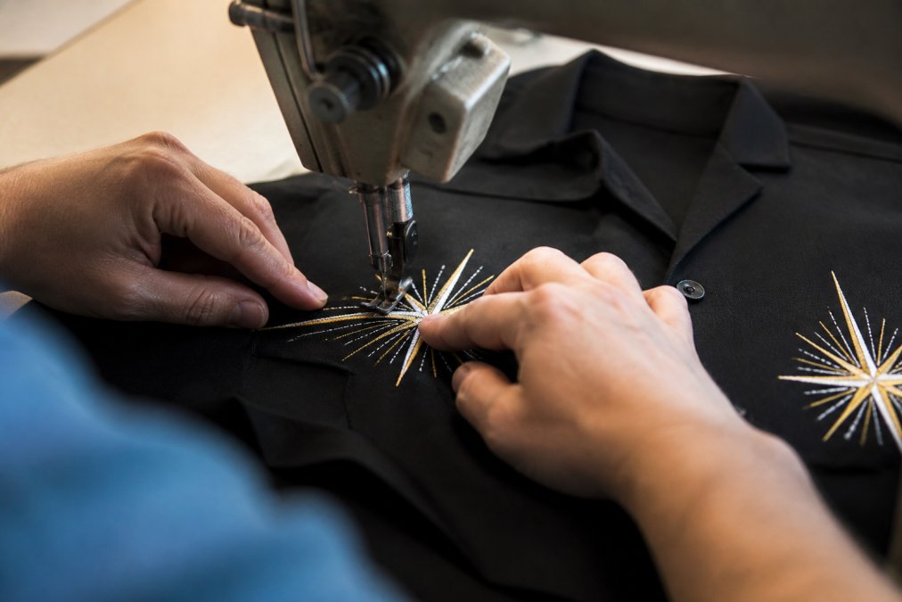 Close-up of skilled hands carefully stitching detailed embroidery onto a black retro bowling shirt, showing intricate custom button work and precise needle placement on quality fabric