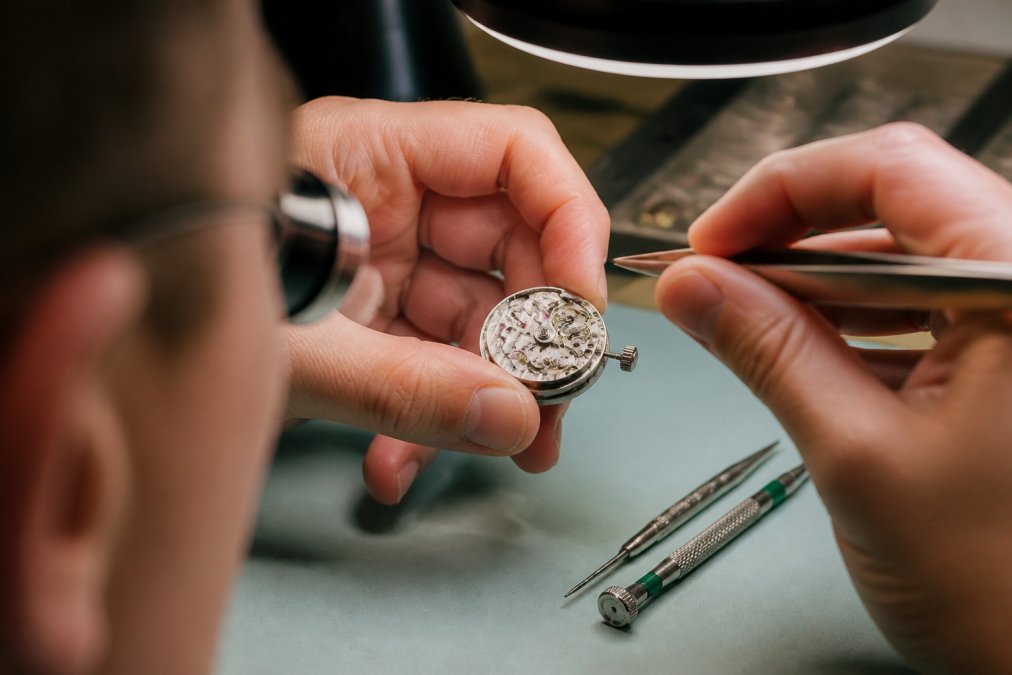 Close-up of watchmaker's hands holding a mechanical movement with visible jewels and bridges under bright magnification lamp, precision tweezers and screwdriver tools visible on the workbench, showing detailed assembly work, showing detailed assembly work