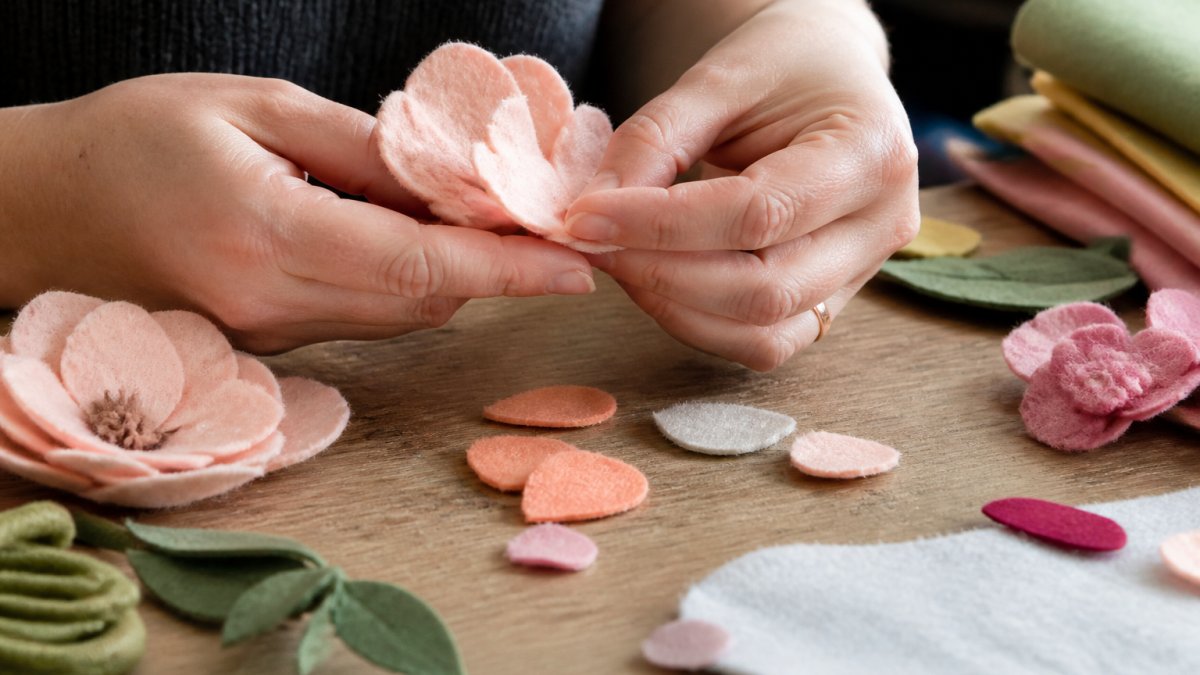 Close-up of hands carefully shaping and arranging individual felt flower petals, showing detailed handcrafting with Merino wool felt in natural studio lighting