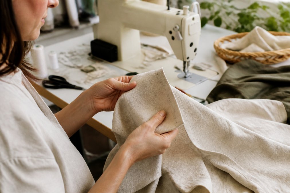 Close-up of a woman's hands feeling the texture of a natural linen blouse, with a sewing machine and fabric scraps visible in the background of a bright studio