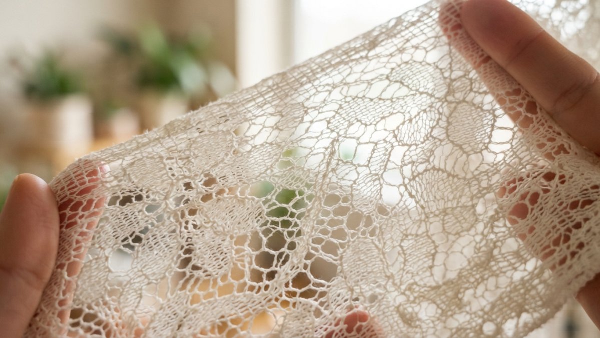 Close-up of delicate lace fabric held up to natural light, showing intricate open-weave pattern with visible gaps that allow light and air to pass through the textile structure