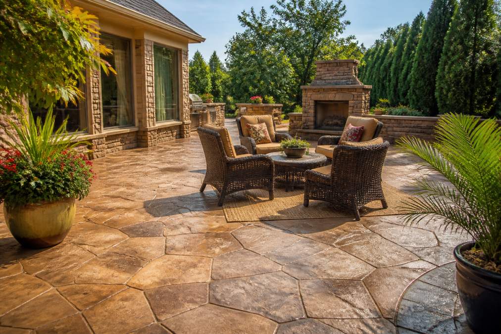 Residential patio with stamped concrete featuring decorative patterns in earth-tone colors, with comfortable outdoor furniture and potted plants creating an inviting outdoor living space under afternoon sunlight