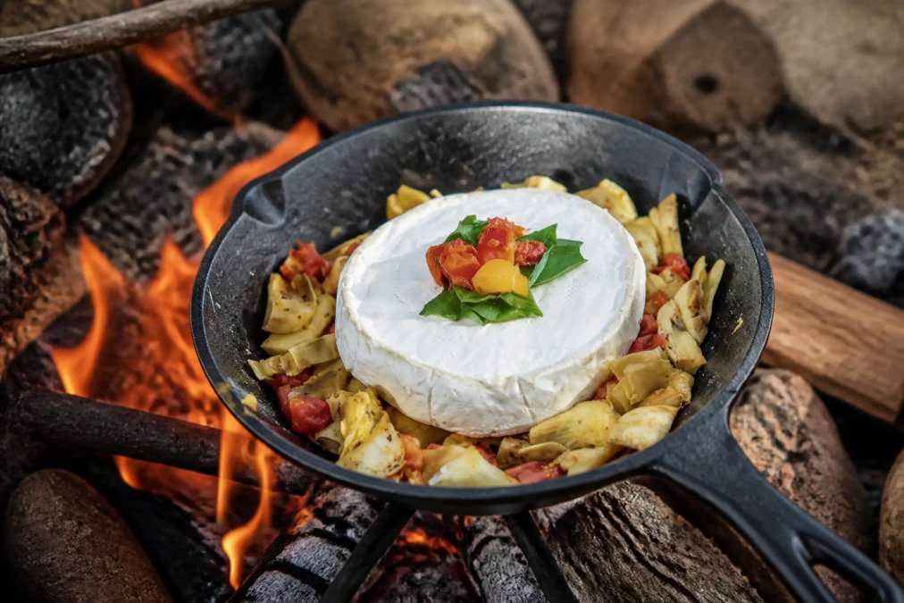 Close-up of a hand forged carbon steel pan sizzling over an open campfire with food cooking, showing the dark patina and even heat distribution across the cooking surface in warm firelight
