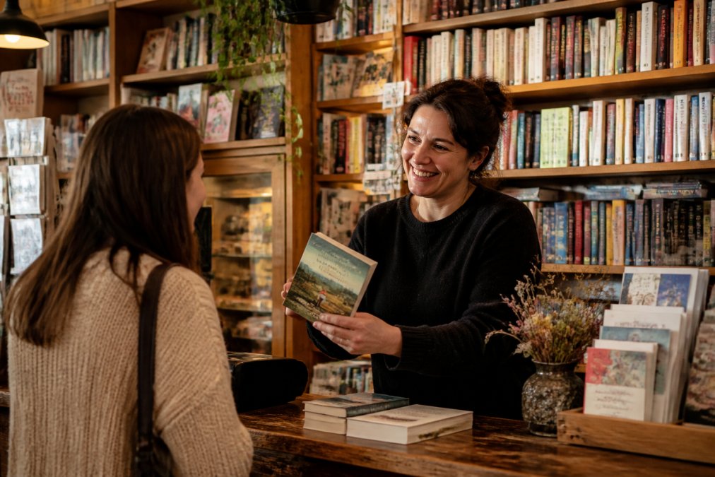 Independent bookshop owner or staff member recommending a book to a customer across the counter, both smiling warmly, in a community-focused retail setting with shelves of books visible in the background