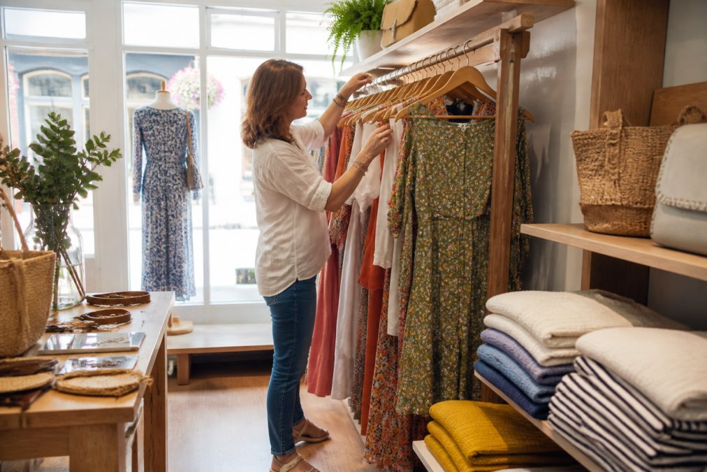 A boutique owner arranging a small display of dresses on a wooden rail in a bright, welcoming shop interior, with folded knitwear and accessories nearby, natural daylight through the window