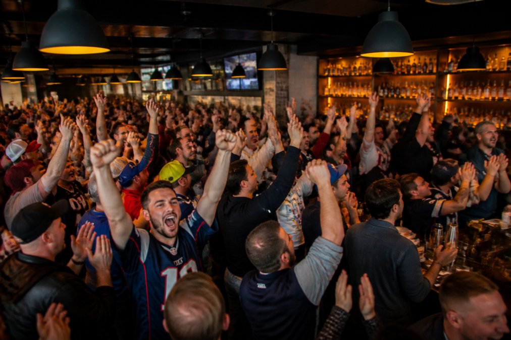 A crowded local sports bar packed with fans cheering at multiple big screens showing a live game, hands raised, vibrant atmosphere with colorful bar lighting