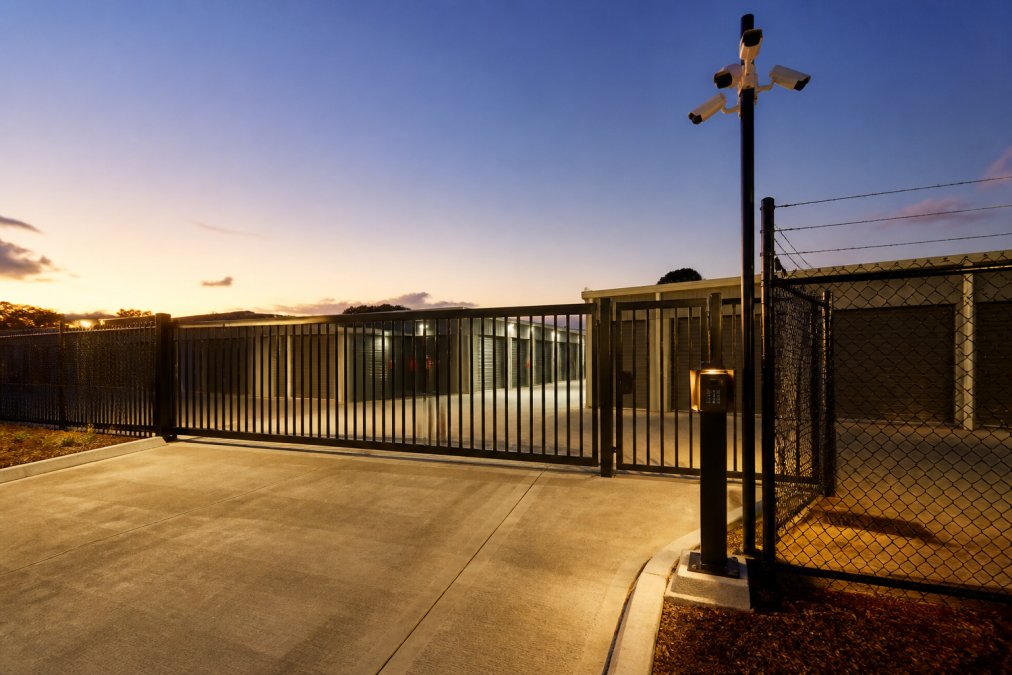 Secure gated entrance to modern storage facility with CCTV cameras mounted on perimeter fence, well-lit access point with coded gate system at dusk