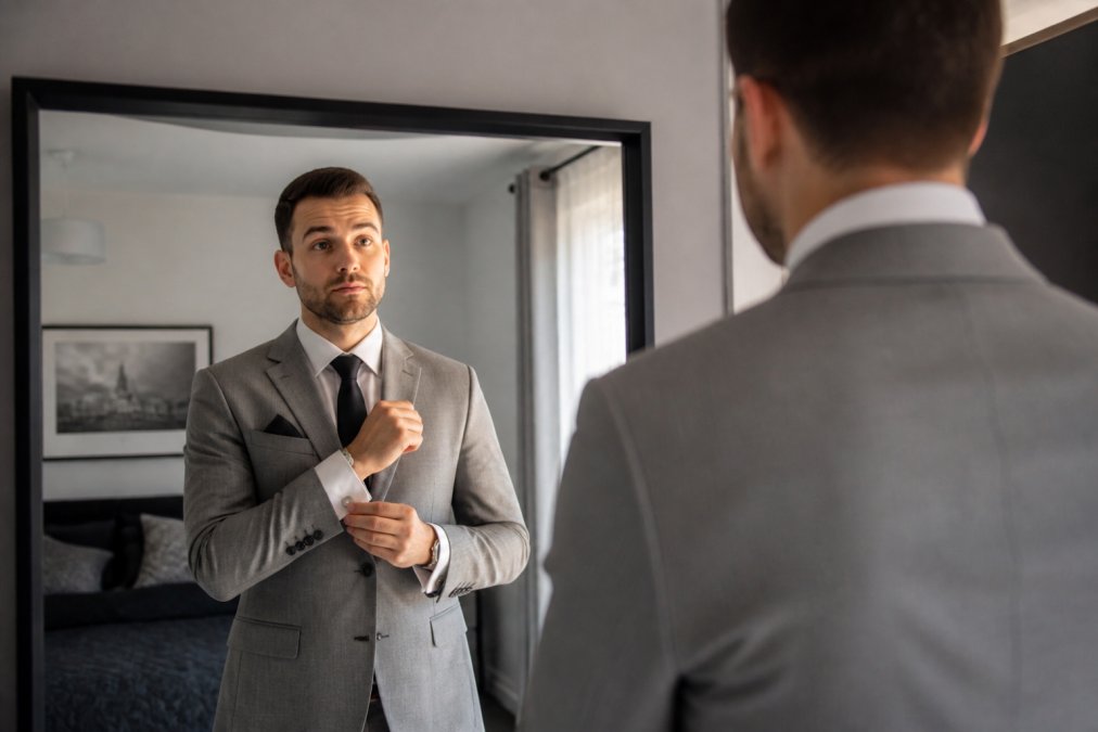 A well-dressed man in a modern, tailored grey suit adjusting his cufflinks while looking in a mirror at home, natural light from a window