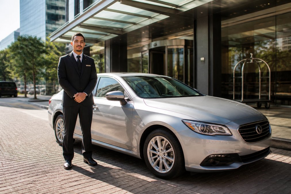 Professional driver in dark uniform standing beside a clean silver sedan outside a modern Perth CBD hotel entrance, morning sunlight