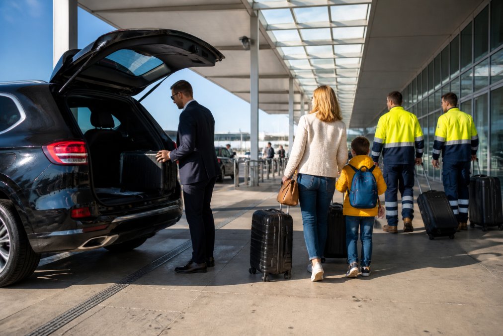 Diverse group of passengers including business professionals, families with children, and FIFO workers boarding a modern black SUV in Perth airport departure area, afternoon light