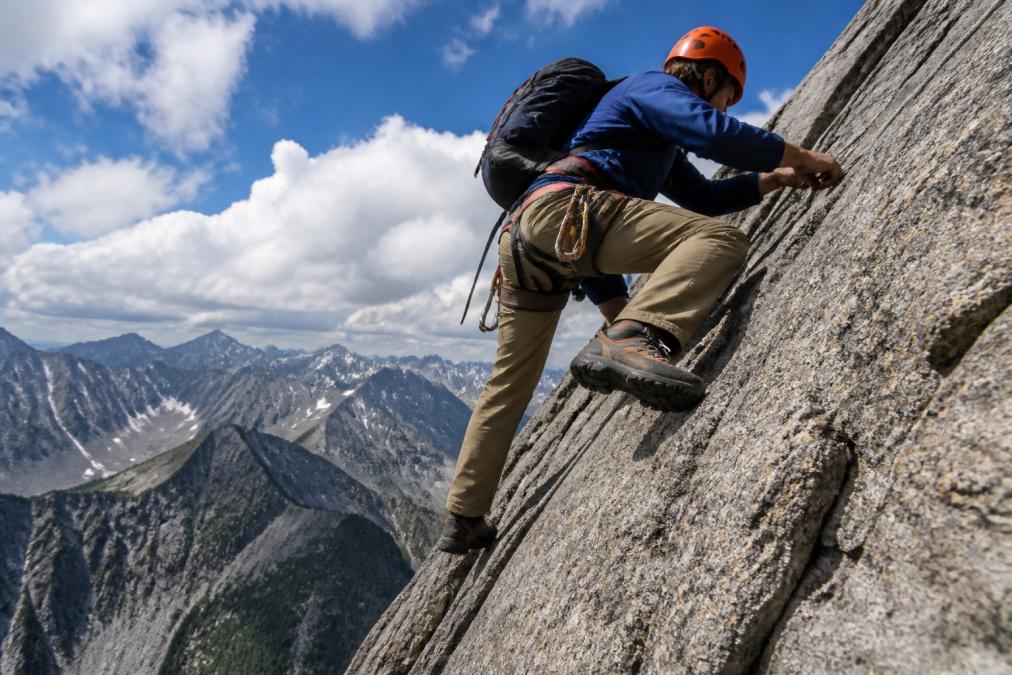 Experienced climber navigating steep rocky mountain terrain wearing professional-grade hiking boots and harness system, demonstrating confident footwork on exposed granite face with dramatic alpine backdrop