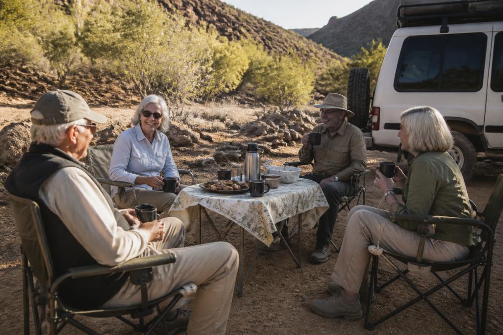 A small group of mature travellers enjoying refreshments and conversation during a scenic break on a 4WD tour, relaxed and unhurried, natural outback lighting with comfortable seating