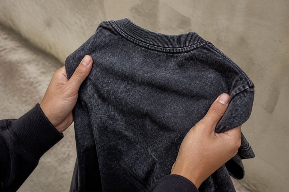 Close-up of hands holding a faded black snow-washed graphic tee, showing the mottled texture and soft wrinkles of the fabric against a neutral urban backdrop