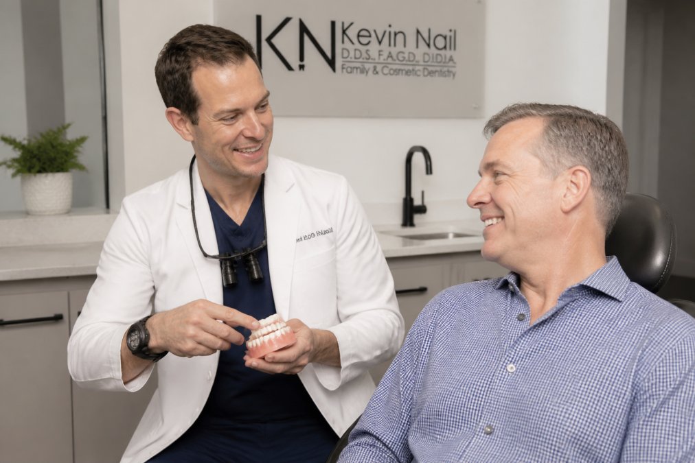 A dentist in a white coat leaning forward to explain a treatment plan to a relaxed middle-aged patient in a modern, bright dental office, both smiling with genuine rapport