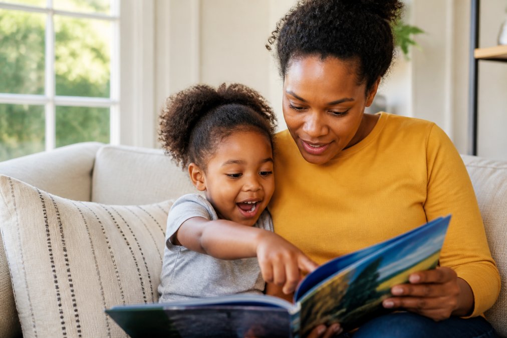 Parent and child sitting together on a couch, reading a colorful adventure book with the child pointing excitedly at the pages, warm natural light streaming from a nearby window