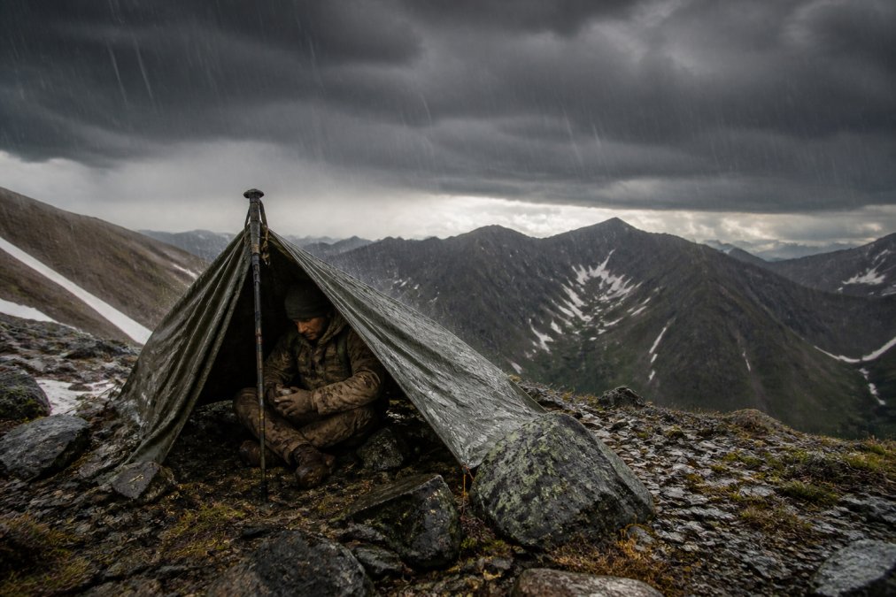A veteran in tactical gear testing a poncho liner shelter in rugged mountainous terrain during heavy rain and wind, with dramatic storm clouds overhead