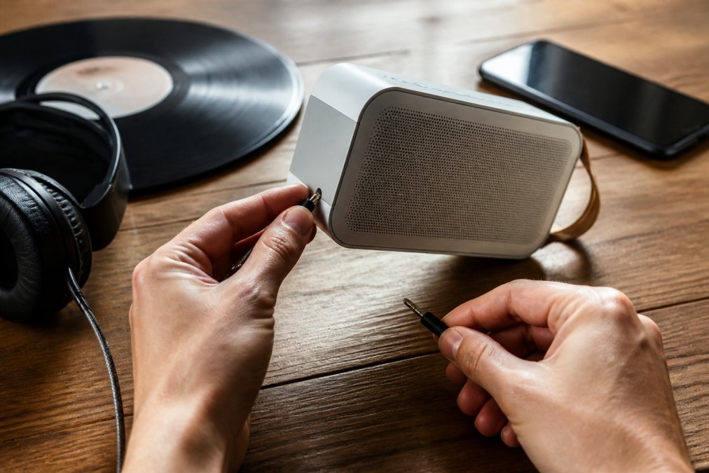 Close-up of hands plugging a wired headphone jack into a portable speaker on a wooden desk, with a smartphone and vinyl record nearby in warm natural light