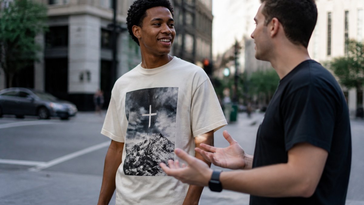 A young adult wearing a graphic faith-based T-shirt standing in a city setting, turning slightly toward a friend who is mid-conversation, natural daylight, candid streetwear look