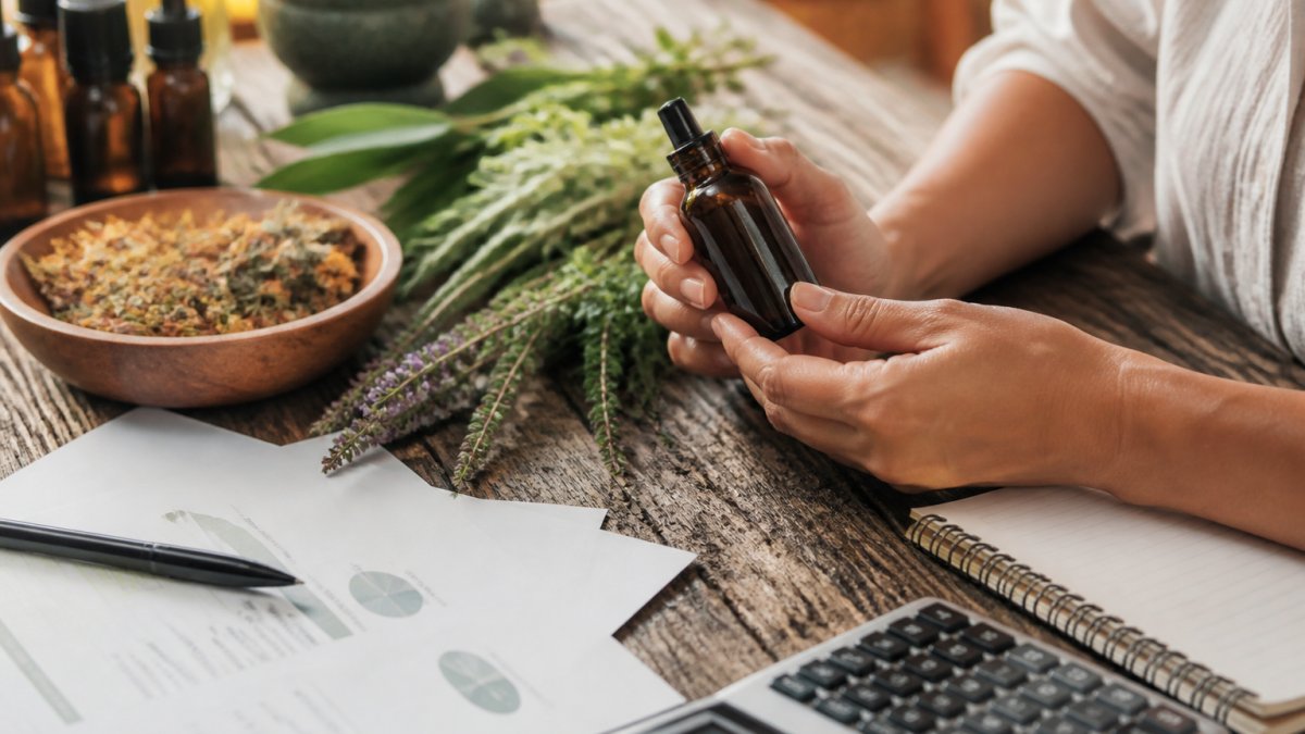 A close-up of hands holding a small amber glass bottle of botanical oil next to fresh herbs and a wooden bowl of dried flowers on a rustic table, with soft natural light