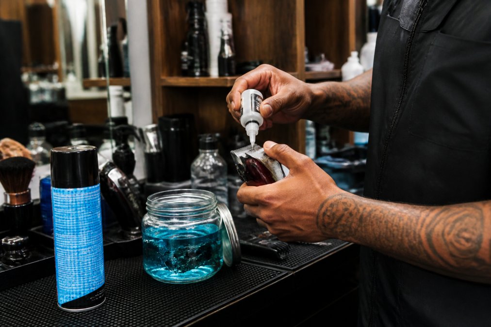 Professional barber's hands applying clipper oil to blade edges over a disinfecting jar with maintenance tools and Andis Cool Care spray visible on the counter