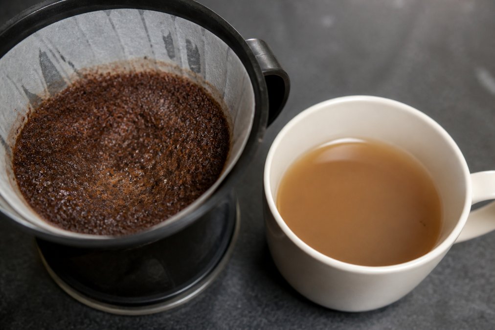 Close-up of wet coffee grounds in a filter basket showing visible particles and moisture, with a cup of pale, thin-looking brewed coffee beside it