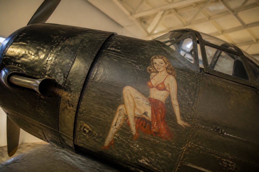 A close-up photograph of a vintage WWII fighter aircraft nose art panel, showing painted pin-up or bomber girl artwork with weathered paint and historical patina, displayed in a museum setting