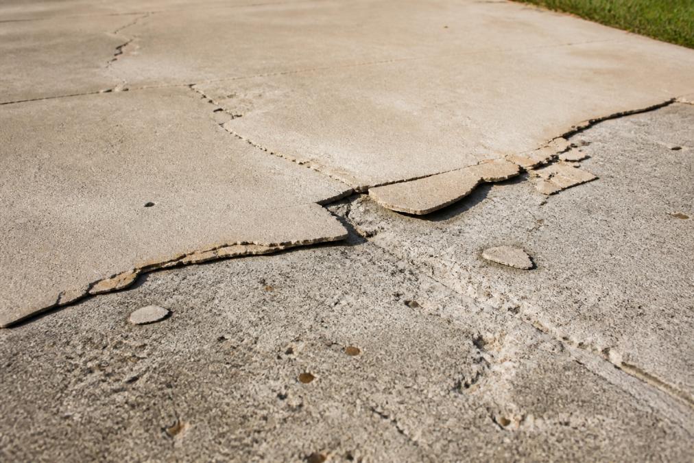 Close-up of a cracked and peeling concrete driveway surface showing visible delamination and spalling, with the failed top coating lifting away from the grey slab underneath, harsh afternoon sunlight emphasising the texture