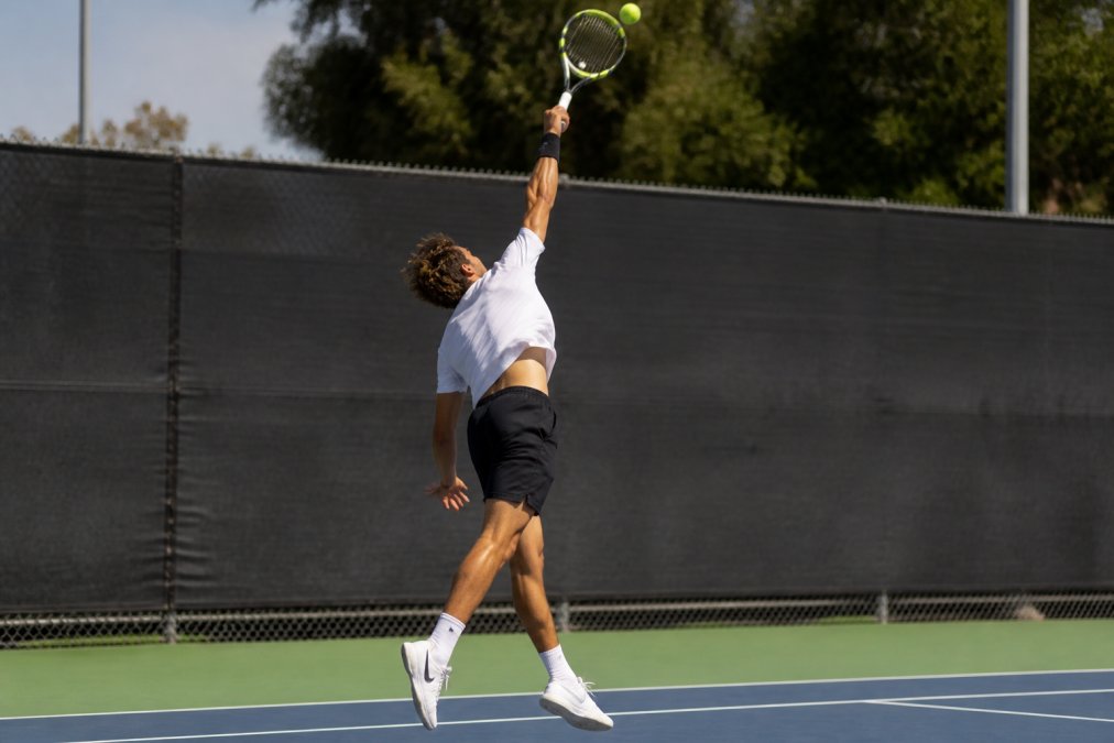 Professional tennis player mid-serve showing proper racket trajectory, weight transfer from back foot to front foot, and full body extension through the kinetic chain in outdoor court lighting