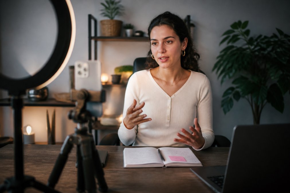 A solo content creator filming a talking-head video in a home studio, smartphone mounted on a tripod in front of a softly glowing ring light, notes visible on a desk beside them