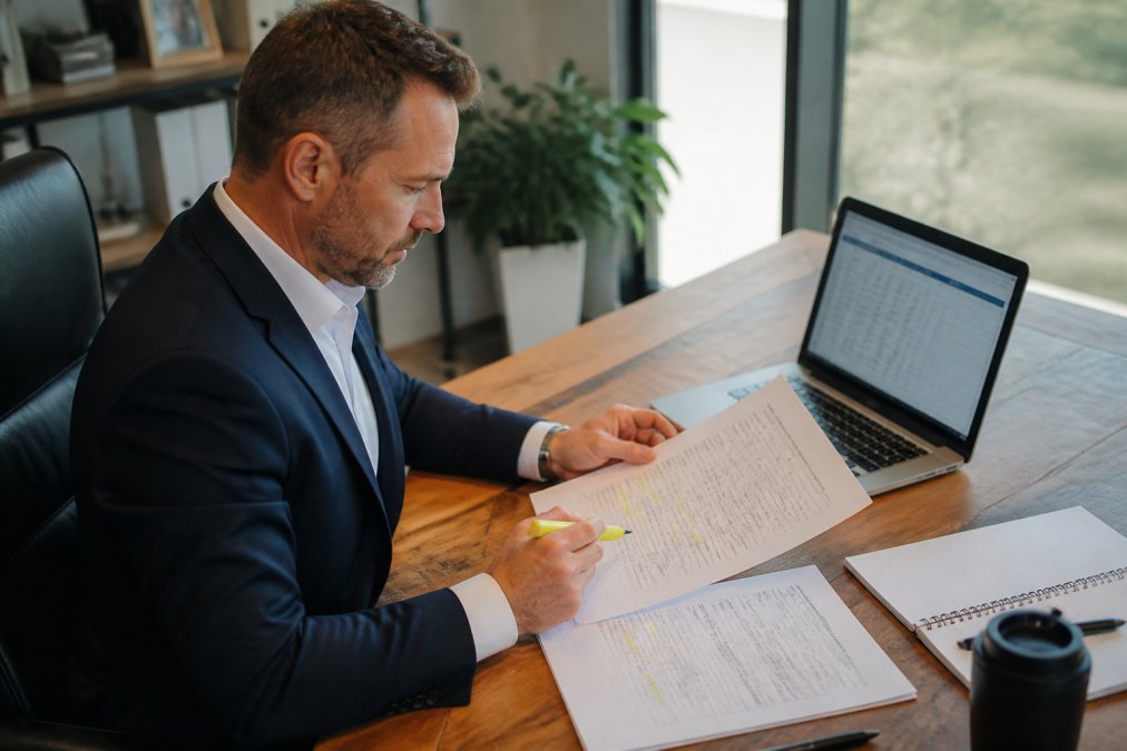Business owner in professional attire sitting at wooden desk, reviewing printed merchant statement with highlighter in hand, laptop open displaying payment processing dashboard with charts, natural window lighting