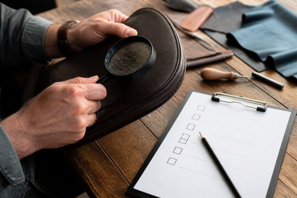 Close-up of hands inspecting the stitching and material of a premium lifestyle product, with a magnifying glass and a checklist on a wooden desk, natural lighting