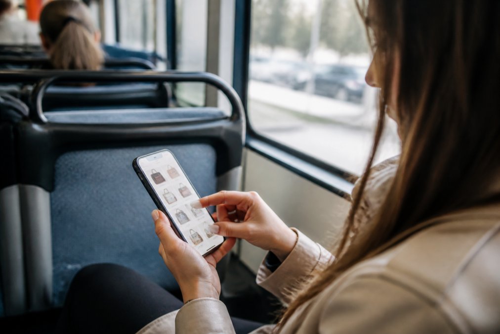 Close-up of hands holding a smartphone with a shopping app open, person browsing trending products while commuting on public transit, natural daylight visible through a window