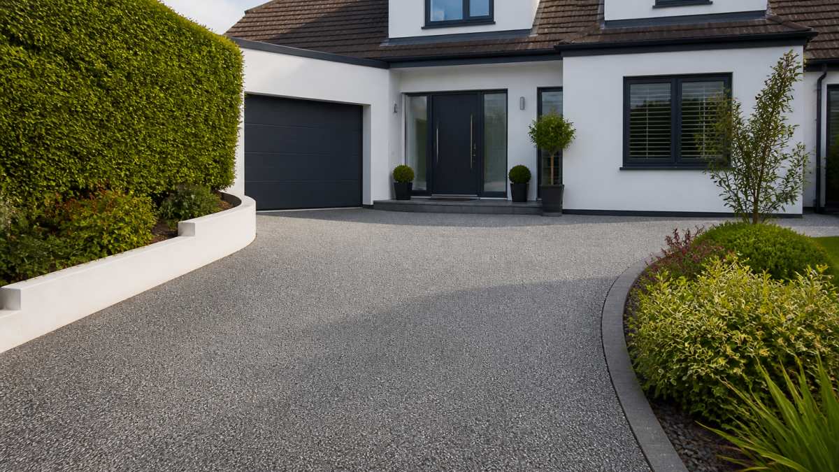 Well-maintained resin bound driveway in neutral grey tone leading to a modern residential property entrance, flanked by manicured borders and contemporary landscaping, photographed in natural daylight