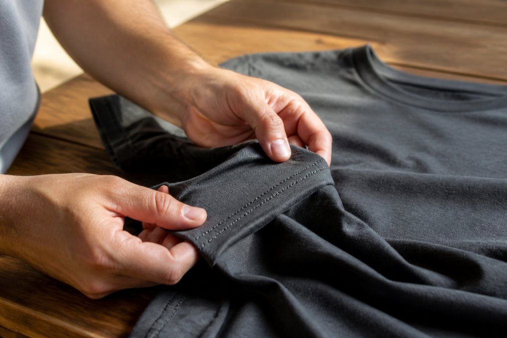 Close-up of hands examining the fabric weave and stitching details of a premium custom t-shirt, showing visible double-stitching at seams and reinforced construction on a wooden table with natural daylight