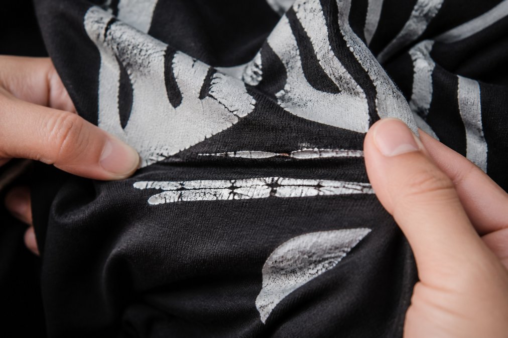 Close-up of hands stretching a black graphic tee fabric, showing the cracked white ink layer separating at a crease under bright overhead lighting