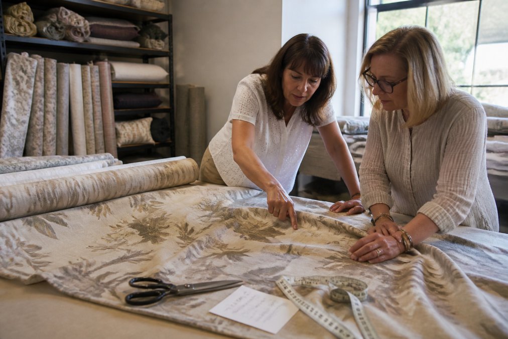 A workroom table with a wide roll of upholstery-weight drapery fabric partly unrolled, fabric shears, a tape measure, and a paper cutting ticket with handwritten measurements, natural window light from the side